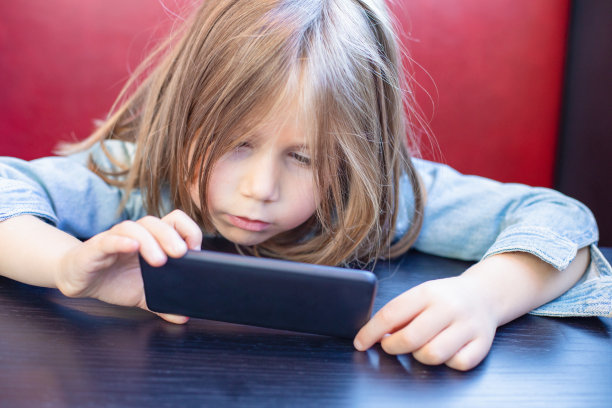 金发小孩坐在红色的背景和黑色的桌子上专注地看智能手机图片下载