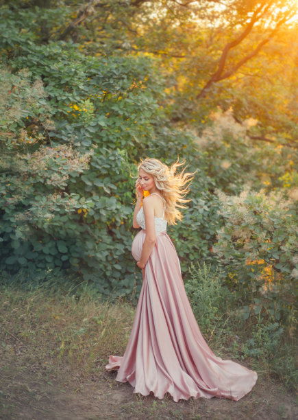 一位年轻的孕妇，头发轻盈飘动，穿着一件粉红色的绸缎豪华长裙，上面有白色的蕾丝花边，微笑着站在森林中的一座小山上，拥抱着自己的肚子。艺术照片图片下载