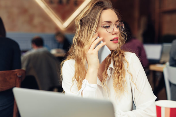 女人戴眼镜，打电话，在咖啡馆用笔记本电脑工作时喝热咖啡。肖像时尚微笑的女人在冬天的衣服喝热咖啡和工作在笔记本电脑。女性冬季风格。——图片图片下载