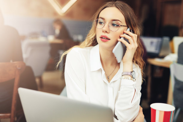 女人戴眼镜，打电话，在咖啡馆用笔记本电脑工作时喝热咖啡。肖像时尚微笑的女人在冬天的衣服喝热咖啡和工作在笔记本电脑。女性冬季风格。——图片图片下载