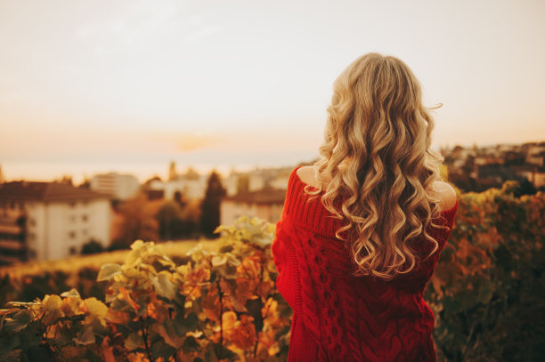户外肖像的女人与金发卷曲的头发欣赏日落，穿着红色套头衫与开放的肩膀，背面的观点。这张照片摄于瑞士洛桑的拉沃葡萄园图片下载