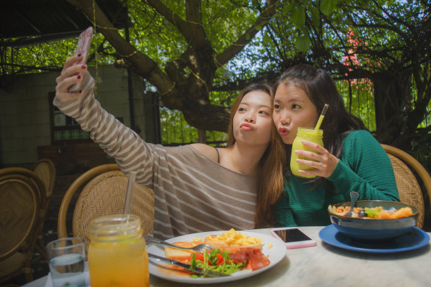 两个年轻快乐和可爱的亚洲中国女友用手机相机自拍照片在网络社交媒体上使用，同时在咖啡厅吃健康的早午餐的生活方式肖像图片下载
