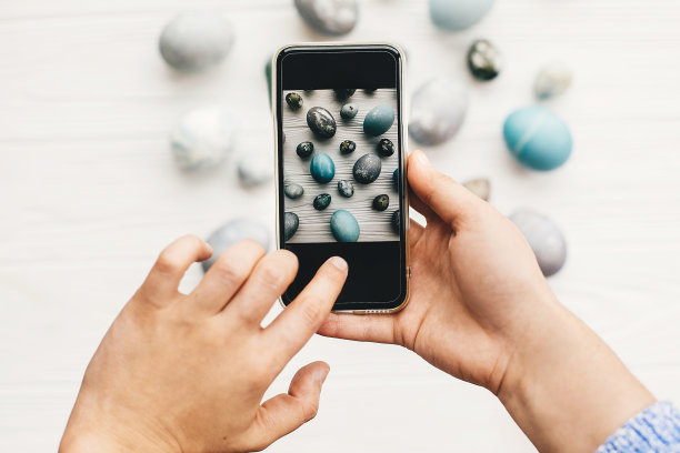 手拿着电话，并使照片时尚的复活节平鸡蛋躺在乡村白色的木头。博客的概念。复活节快乐。现代的复活节彩蛋用天然染料涂成灰色图片下载