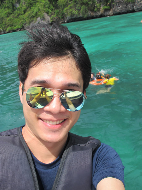 亚洲英俊的男子在泳装与救生衣微笑，并采取了在美丽的水中央的一艘船自拍照片。图片下载