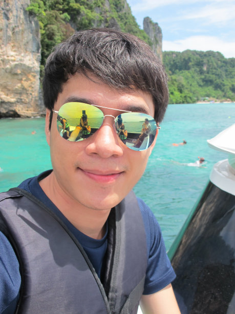 亚洲英俊的男子在泳装与救生衣微笑，并采取了在美丽的水中央的一艘船自拍照片。图片下载