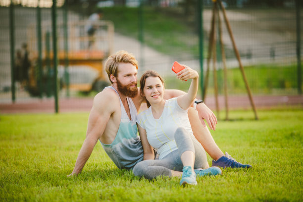 运动和技术。年轻相爱的白人情侣在户外公园草坪上锻炼后休息，在日落时分用智能手机自拍图片下载