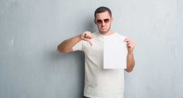 年轻的白人男子在灰色垃圾墙持空白纸与愤怒的脸，消极的迹象显示不喜欢拇指向下，拒绝概念图片下载