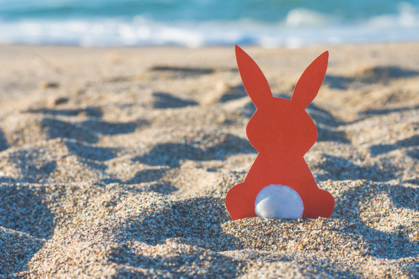 创意复活节照片的红纸兔子在沙滩上的沙滩上日落。热带国家庆祝复活节的概念。图片下载
