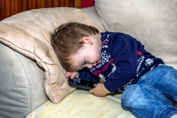 漂亮的小男孩穿着圣诞毛衣躺在客厅的沙发上玩着智能手机图片下载