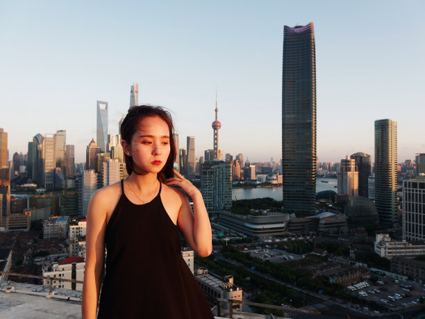 美丽的年轻女子在性感的黑色连衣裙摆姿势在大厦屋顶上与模糊的上海外滩地标建筑背景黄昏的光。情感、人、美、生活理念。图片下载