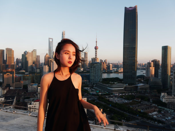 美丽的年轻女子在性感的黑色连衣裙摆姿势在大厦屋顶上与模糊的上海外滩地标建筑背景黄昏的光。情感、人、美、生活理念。图片下载