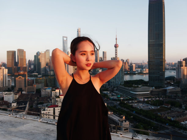 美丽的年轻女子在性感的黑色连衣裙摆姿势在大厦屋顶上与模糊的上海外滩地标建筑背景黄昏的光。情感、人、美、生活理念。图片下载