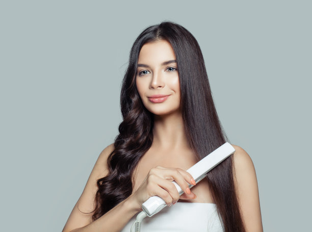 用直发器拉直长发和卷发的漂亮女人。可爱的微笑女孩直发健康的黑发与扁铁。头发问题与护发理念图片下载