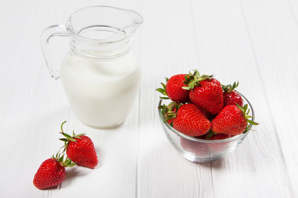 新鲜的草莓和一壶牛奶，白色的木背景上的一个碗里装着红色的草莓。几个单独的浆果。水平的照片图片下载