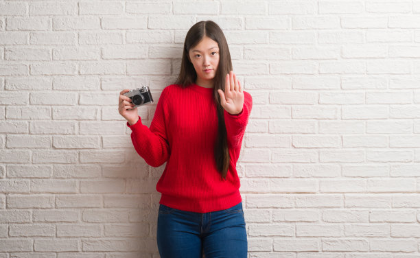 一名年轻的中国女子越过砖墙，手举老式相机，做着严肃自信的手势和防御手势图片下载