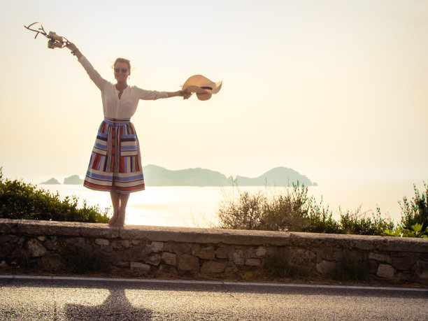 快乐的年轻女子戴着帽子，穿着优雅多彩的裙子，赤脚站在海边的一面墙在夕阳意大利庞扎岛。武器扩散。图片下载