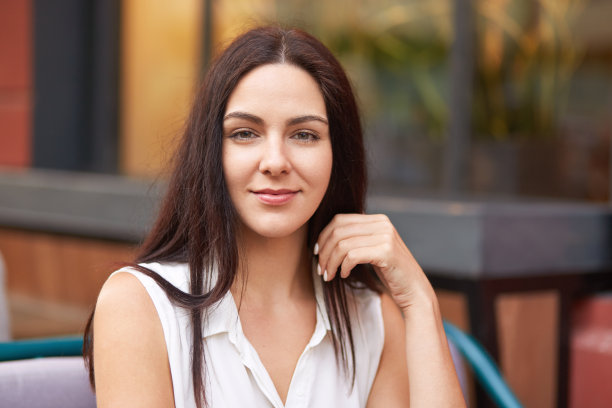 照片中美丽的黑发女士，淡妆，健康的皮肤，在镜头前看起来很直接，穿着白色衬衫，在户外餐厅模糊的背景下摆姿势。美丽的概念。图片下载