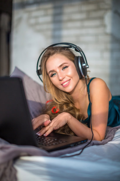 一个女人躺在床上，拿着笔记本电脑和耳机，交叉着双腿放松着，微笑着图片下载