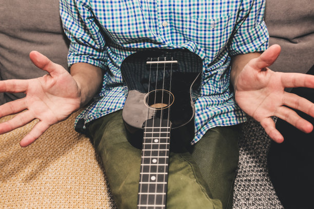 一个穿着格子衬衫的男人正在家里弹尤克里里吉他。大家的手在弦上拨动和弦。音乐创造生活理念。每个人的业余爱好。有趣的吉他照片。图片下载
