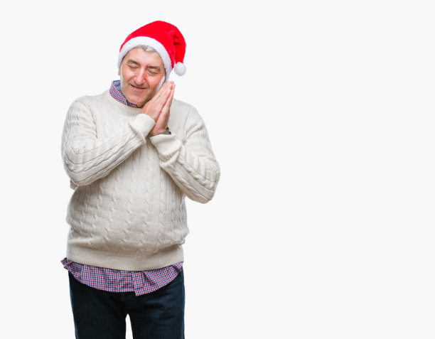 英俊的老男人戴着圣诞帽在孤立的背景睡累了，做梦，摆姿势与手在一起，而闭着眼睛微笑。图片下载