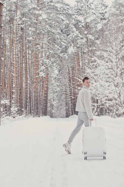 一个年轻的时尚男子在雪地上举着手提箱，竖起大拇指。寒假旅游概念。图片下载