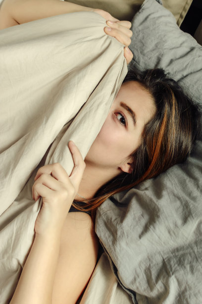 一个躺在床上不想醒来的年轻女人的肖像。快活的女孩躺在床上，把脸藏在被子里。垂直的照片。图片下载