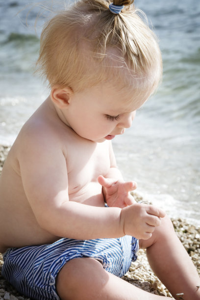 一个可爱的金发婴儿的照片在卵石海滩附近的海边，暑假在海边图片下载