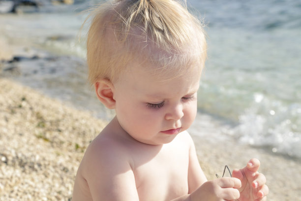 一个可爱的金发婴儿的照片在卵石海滩附近的海边，暑假在海边图片下载