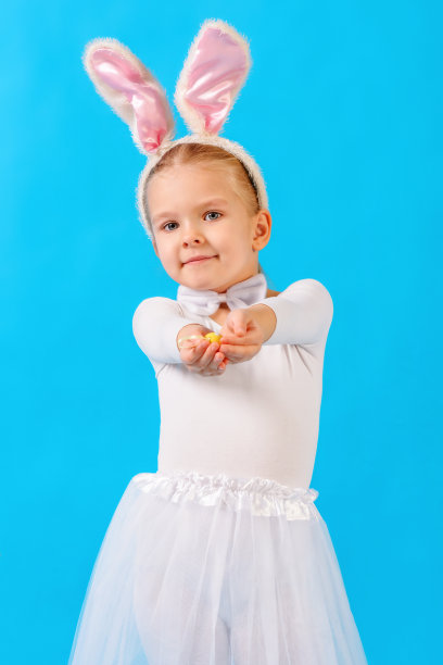 穿着白兔服装的小女孩。那孩子手里拿着一个复活节彩蛋。可爱的兔子，节日的象征。明亮的照片上的蓝色背景。图片下载