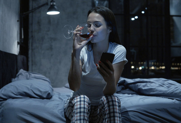 年轻女性晚上在卧室里一边喝酒一边用智能手机交流图片下载