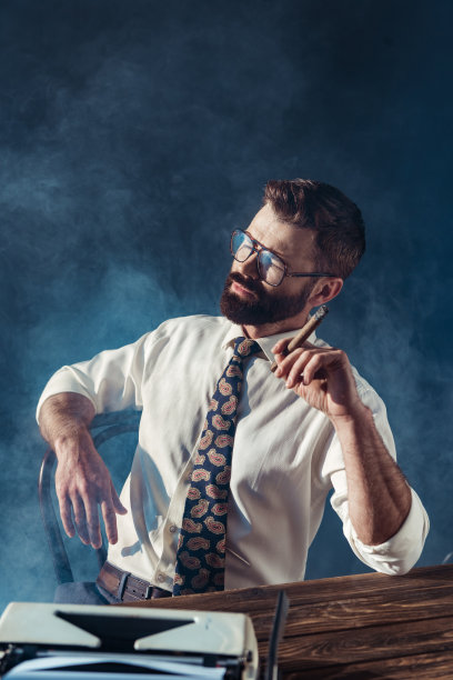 深思熟虑的大胡子记者坐在桌子前，桌上放着老式打字机，灰色的背景上吸烟图片下载