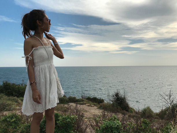 身穿白衣的女子站在山上，望着日落和蓝天下的大海图片下载