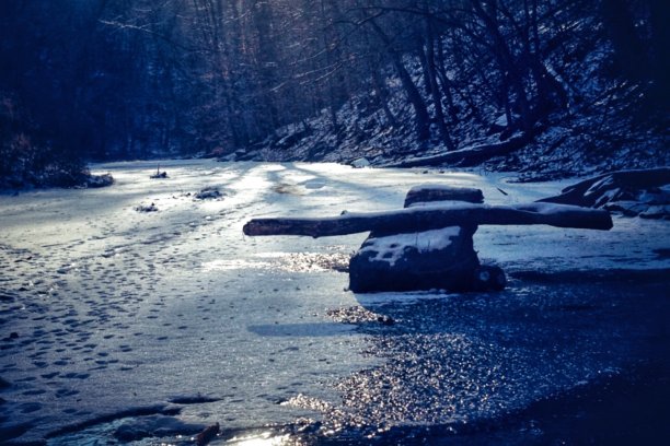 黄昏时分，一根倒下来的圆木落在冰雪覆盖的小溪中间的一块岩石上图片下载