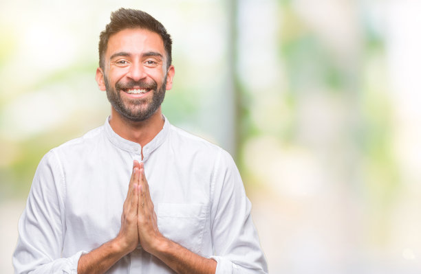 在孤立背景下，双手合十祈祷的西班牙裔成年男子，请求原谅，面带自信的微笑图片下载