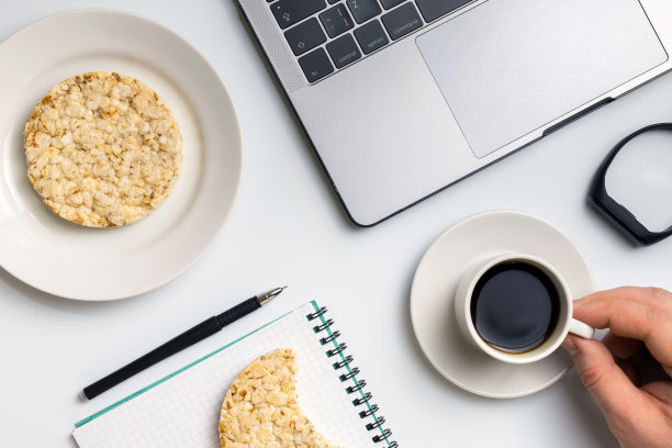 在工作休息时间吃健康的零食。运动员一边吃着锅巴，一边喝着咖啡，旁边是笔记本和笔记本。白色有组织的桌子。图片下载