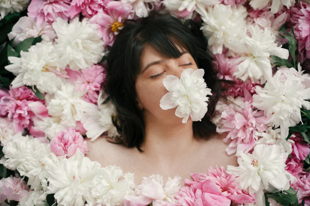 一幅波西米亚女子的画像，她嘴里叼着白色的牡丹，躺在粉红色的牡丹中。创意艺术花卉照片。芳香和水疗概念。国际妇女日。美丽的黑发女孩在许多花图片下载