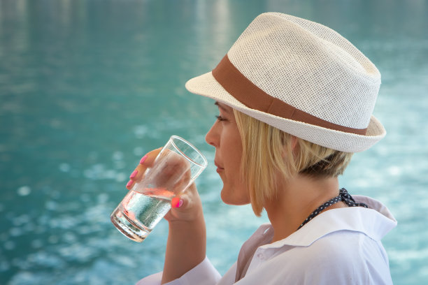 在绿松石般的大海背景下，一个金发白种人女孩从一个汗津津的杯子里喝着冷水图片下载