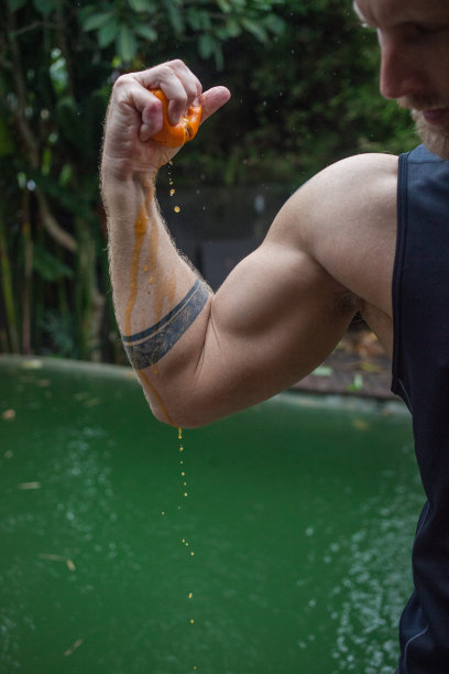 照片中，一名男子在泳池边捏着一个橙子，橙汁在他的手臂上流淌，他身上有纹身，他的肱二头肌和肩膀都很强壮，展示了一种充满水果和蔬菜的健康生活方式图片下载