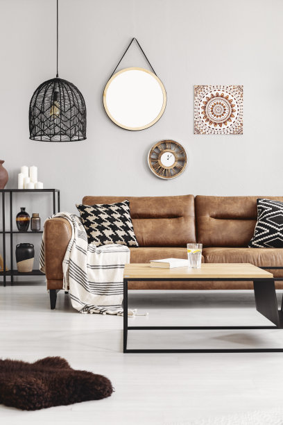 棕色的皮沙发，毯子和图案的坐垫，明亮的客厅内部的真实照片，咖啡桌，柠檬水，书和镜子，墙上的钟和海报图片下载