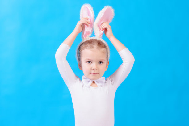 蓝色背景下穿着白兔服装的小女孩。一个小孩摸了摸他的耳朵。美丽的照片与空间的文字和广告。图片下载