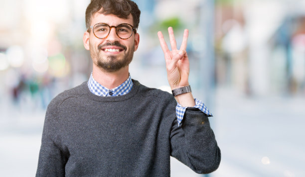 年轻英俊聪明的男人戴着眼镜在孤立的背景显示和指向第三个手指，同时微笑着自信和快乐。图片下载