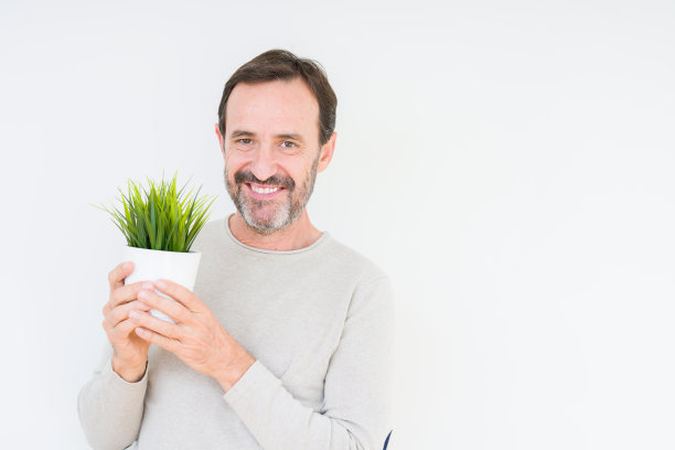 老人抱着绿色植物在孤立的背景与快乐的脸站和微笑与自信的微笑显示牙齿图片下载