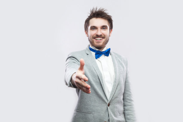 肖像快乐牙齿微笑英俊的胡子男人在休闲灰色西装，蓝色领结站着，看和给予手问候或握手图片下载