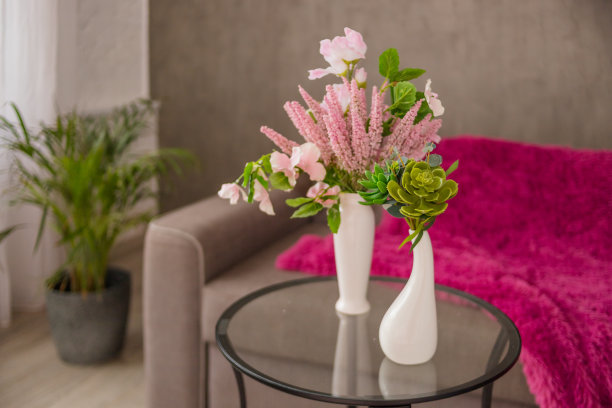 在时尚的客厅里，圆形玻璃咖啡桌上放着两个白色花瓶，配上灰色的斯堪的纳维亚沙发。现代明亮的室内。花瓶植物，花，肉质植物和棕榈树在花盆的背景。家居装饰室内。图片下载