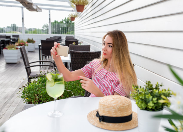 夏日咖啡馆里的美女。在手的智能手机铃声与耳机。照片和观看互联网视频。视频通信，通过社交网络呼叫互联网应用。图片下载