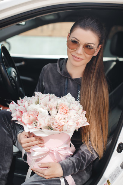 一个漂亮的年轻女子，坐在车里的驾驶座上拿着一个粉红色的盒子里面装满了温柔的，白色的，带着粉红花边的郁金香花图片下载