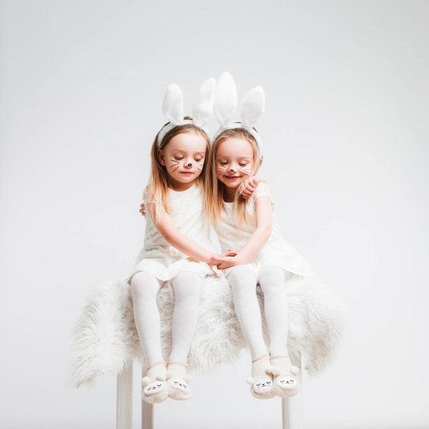 一对金发的双胞胎，穿着白裙子，长着兔耳朵。灰色背景上的工作室照片。孩子们庆祝复活节。图片下载