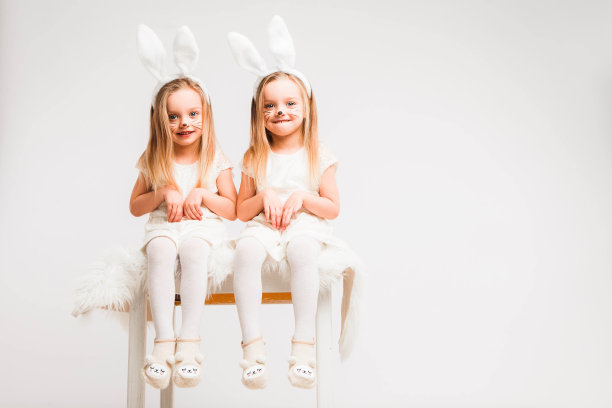 一对金发的双胞胎，穿着白裙子，长着兔耳朵。灰色背景上的工作室照片。孩子们庆祝复活节。图片下载