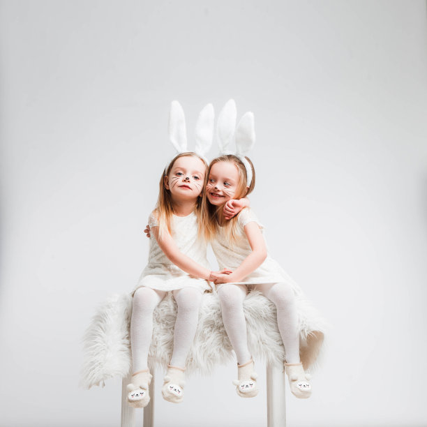 一对金发的双胞胎，穿着白裙子，长着兔耳朵。灰色背景上的工作室照片。孩子们庆祝复活节。图片下载