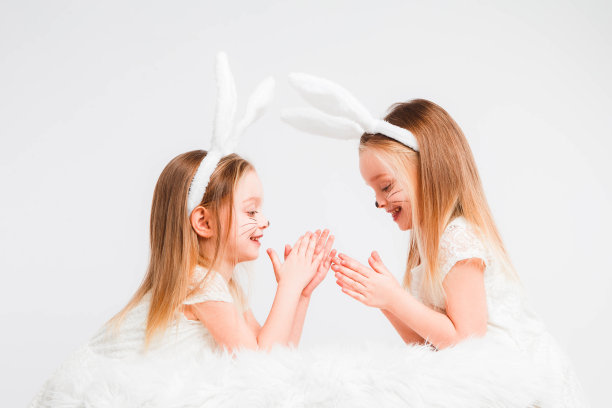 一对金发的双胞胎，穿着白裙子，长着兔耳朵。灰色背景上的工作室照片。孩子们庆祝复活节。图片下载